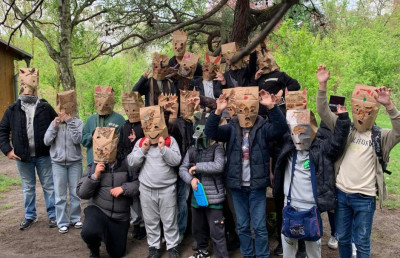 Gruppe von Kindern mit selbstgebastelten Papiermasken verschiedener Gesichter unter einem Baum