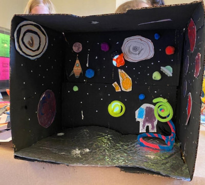 Selbstgebasteltes Weltraum-Diorama in einem schwarzen Schuhkarton mit Planeten, Raketen und Sternen