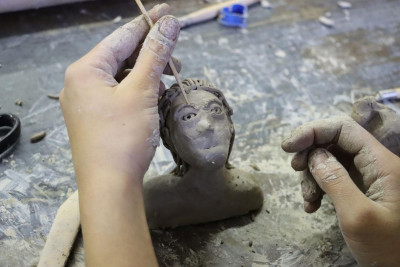 Hände formen mit einem Werkzeug das Gesicht einer kleinen Tonfigur in einer Keramikwerkstatt