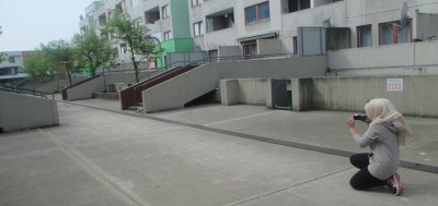 Person fotografiert kniend auf einem Betonplatz vor Wohngebäuden